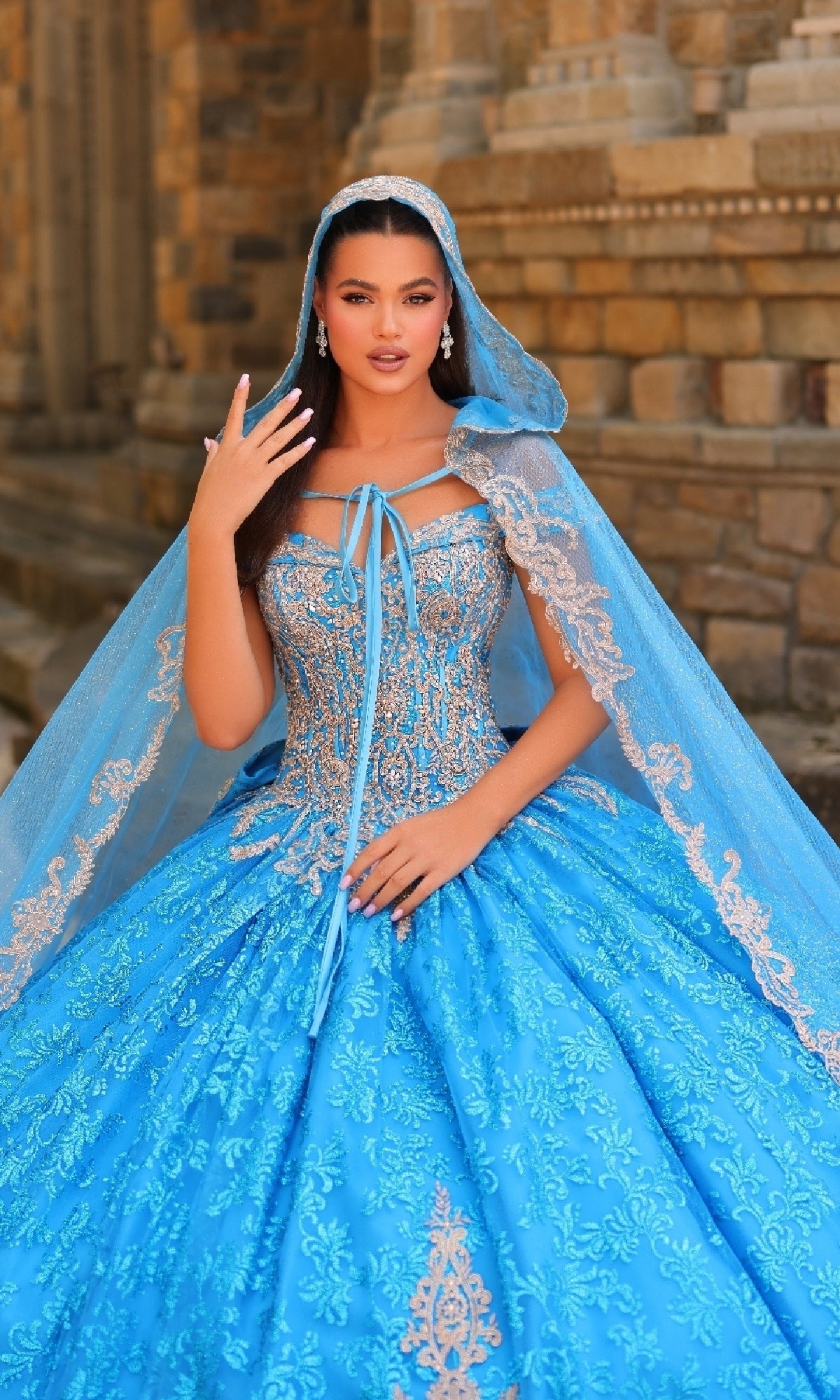 A woman wearing the Quinceanera Dress 54324 By Amarra, featuring ornate blue lace embroidery and a matching lace-trimmed cape, stands in front of a stone building.